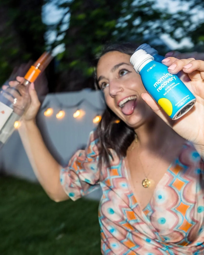 More Labs Morning Recovery bottle held up at an outdoor gathering with a bottle of liquor in the background.