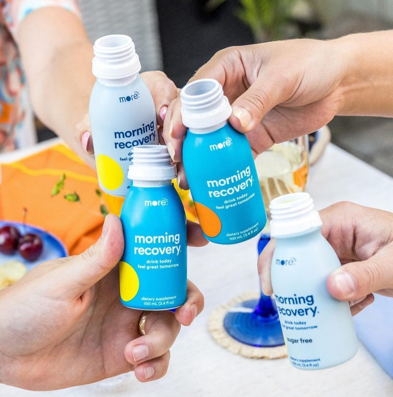People holding and clinking More Labs Morning Recovery bottles together at a table.