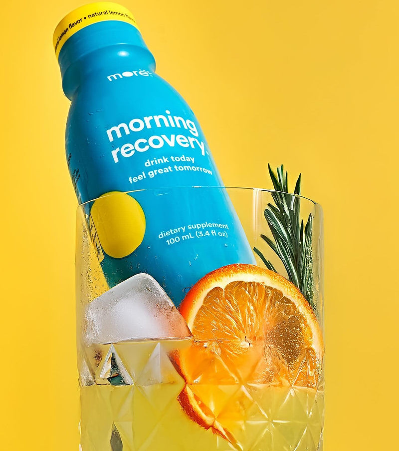 More Labs Morning Recovery dietary supplement in a glass with ice, an orange slice, and rosemary on yellow.