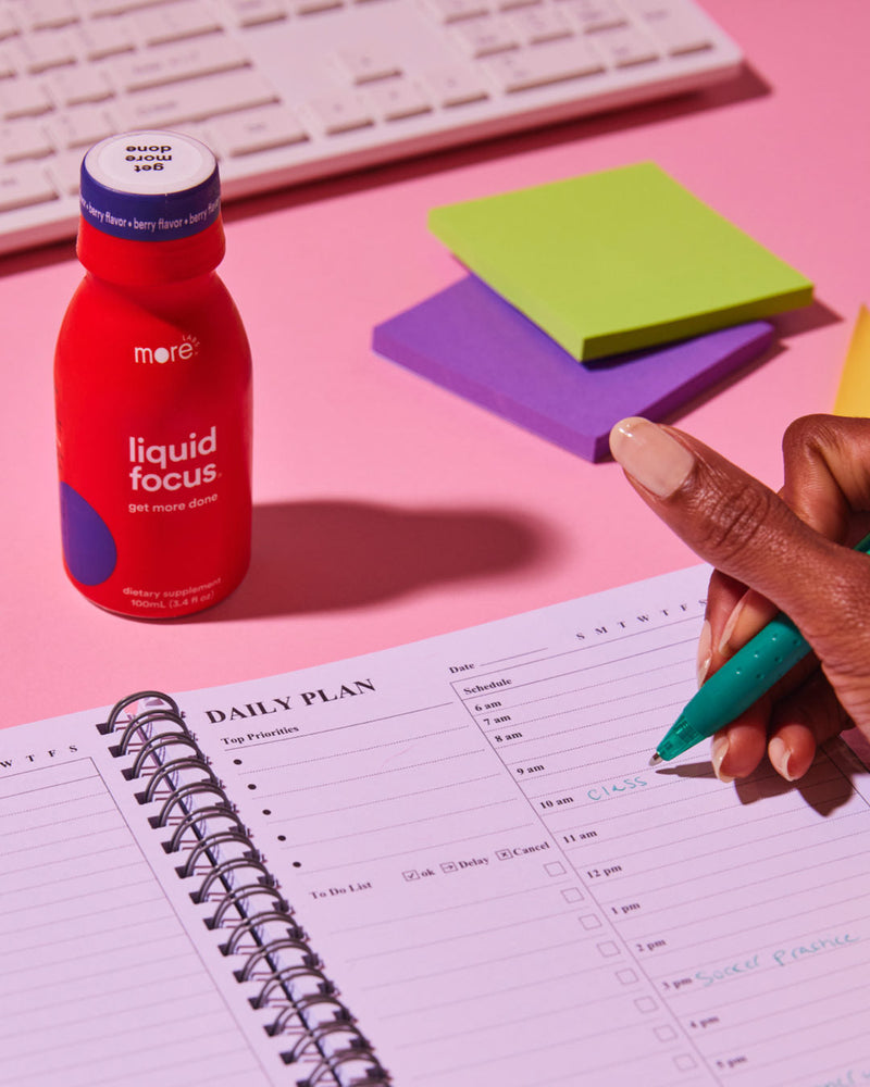 More Labs Liquid Focus bottle on a desk beside a planner and pen as a hand writes a daily schedule.
