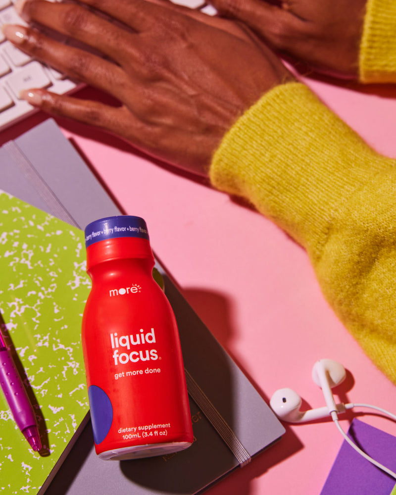 A woman typing on a keyboard with a Liquid Focus bottle and earphones beside her hands.