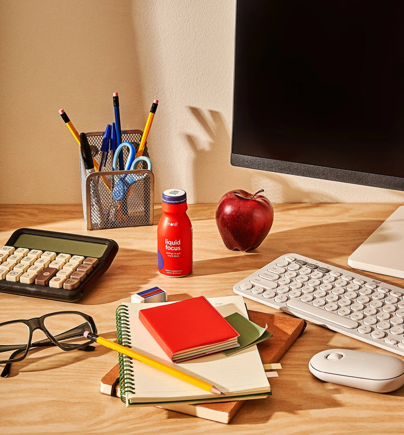 More Labs Liquid Focus bottle on a work desk with notebooks, keyboard, calculator, pencils, and an apple.