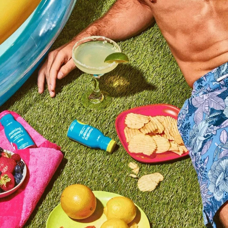 More Labs Morning Recovery bottles beside a drink, chips, and citrus on the grass.