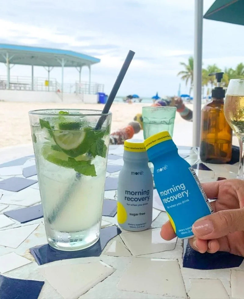 More Labs Morning Recovery bottles beside a mint and lime drink at a beachfront table.