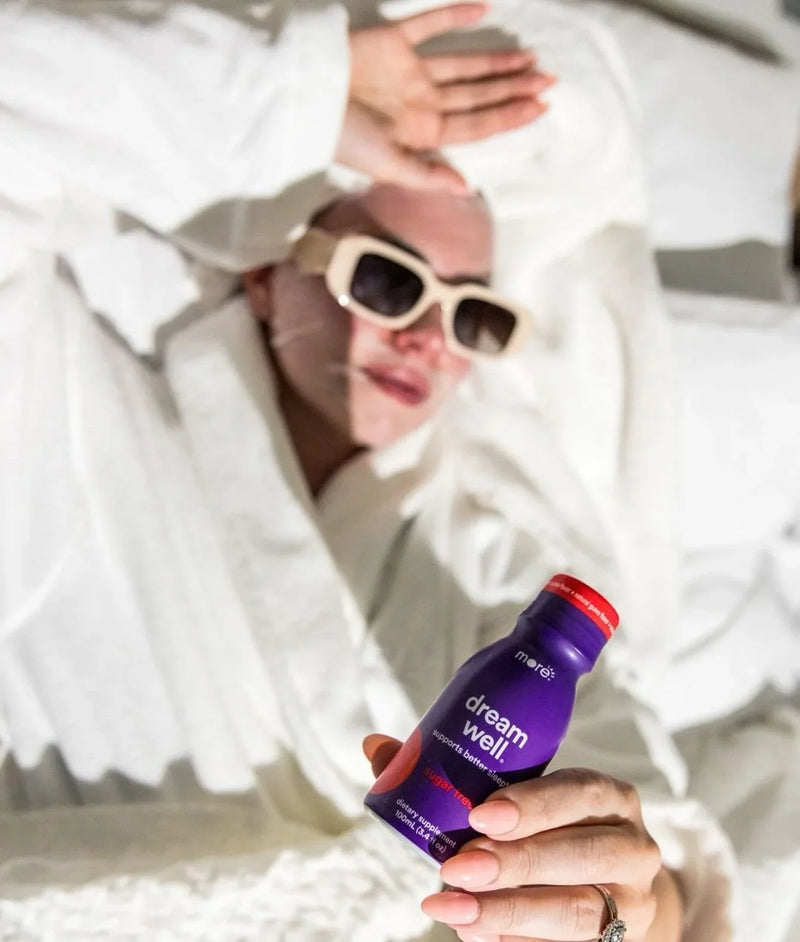 Person relaxing in bed holds a More Labs Dream Well bottle while wearing sunglasses.