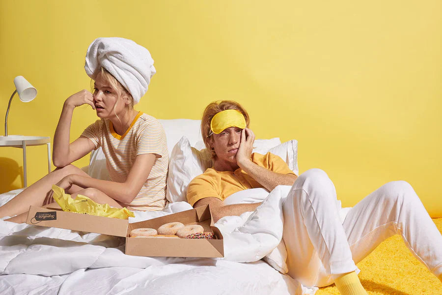 A man and a woman sitting in bed, looking tired, wearing casual clothes, with donuts on a tray and a yellow background.