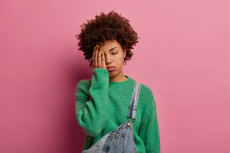 A tired woman covering one eye on a pink background.