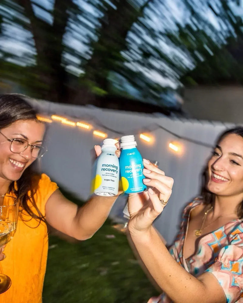 Two women toast with More Labs Morning Recovery bottles at an outdoor gathering.