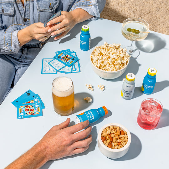 Friends playing cards around a table with Morning Recovery bottles, drinks, popcorn, and snacks during a game night.