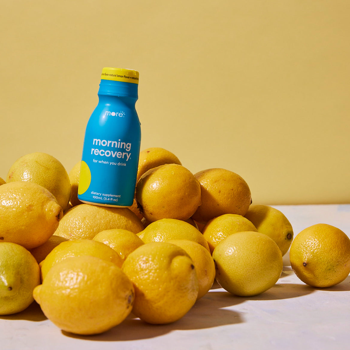 Blue Morning Recovery lemon flavor bottle placed among fresh lemons on a soft yellow background, highlighting the citrus-inspired wellness drink product.
