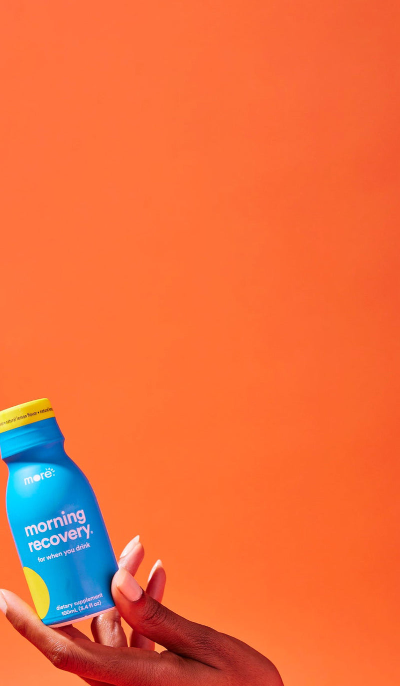 Hand holding a blue Morning Recovery bottle with a yellow cap against a vibrant orange background, showcasing the drink product in a bold lifestyle-style promotional image.
