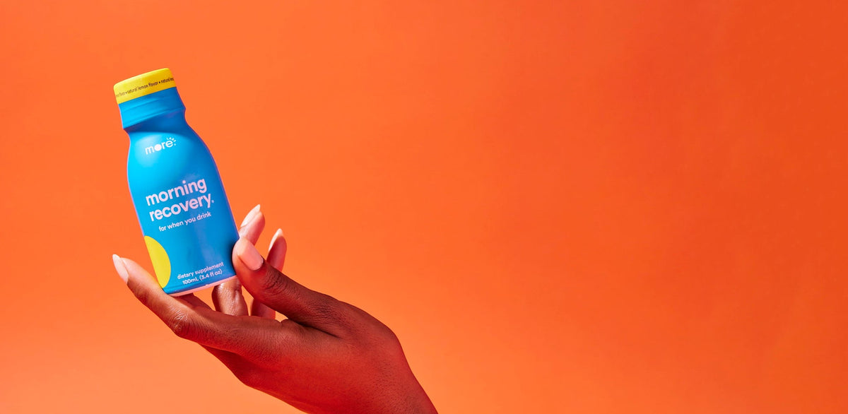 Hand holding a blue Morning Recovery bottle with a yellow cap against a vibrant orange background, showcasing the drink product in a bold lifestyle-style promotional image.
