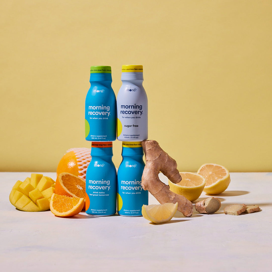 Four Morning Recovery wellness shot bottles displayed with fresh citrus fruits and ginger on a light yellow background, featuring original and sugar-free varieties.
