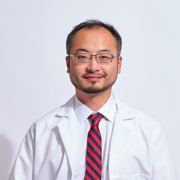 Portrait of a medical professional wearing a white lab coat, glasses, and a red striped tie against a light gray background.
